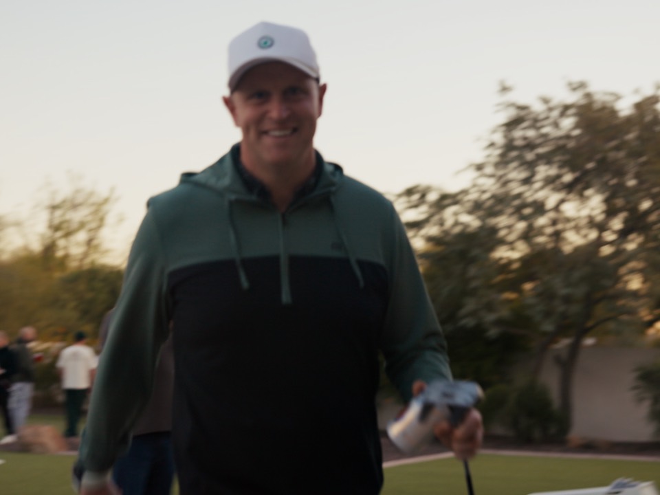 A guest walking up grinning with a drink, friends behind at the house